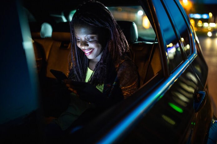 African woman driving on back seat in a car at night and using phone